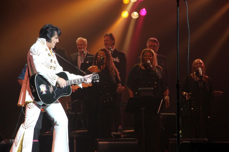 A tribute act dressed as Elvis wearing a white jumpsuit, plays a guitar.